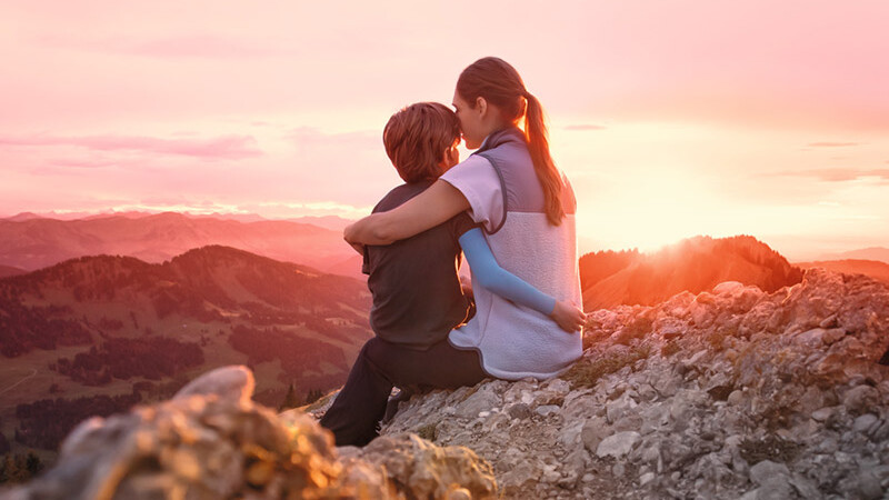 En mamma sitter med sitt barn på en bergstopp. Barnet bär en kompressionsärm. En mamma sitter med sitt barn på en bergstopp. Barnet bär en kompressionsärm.