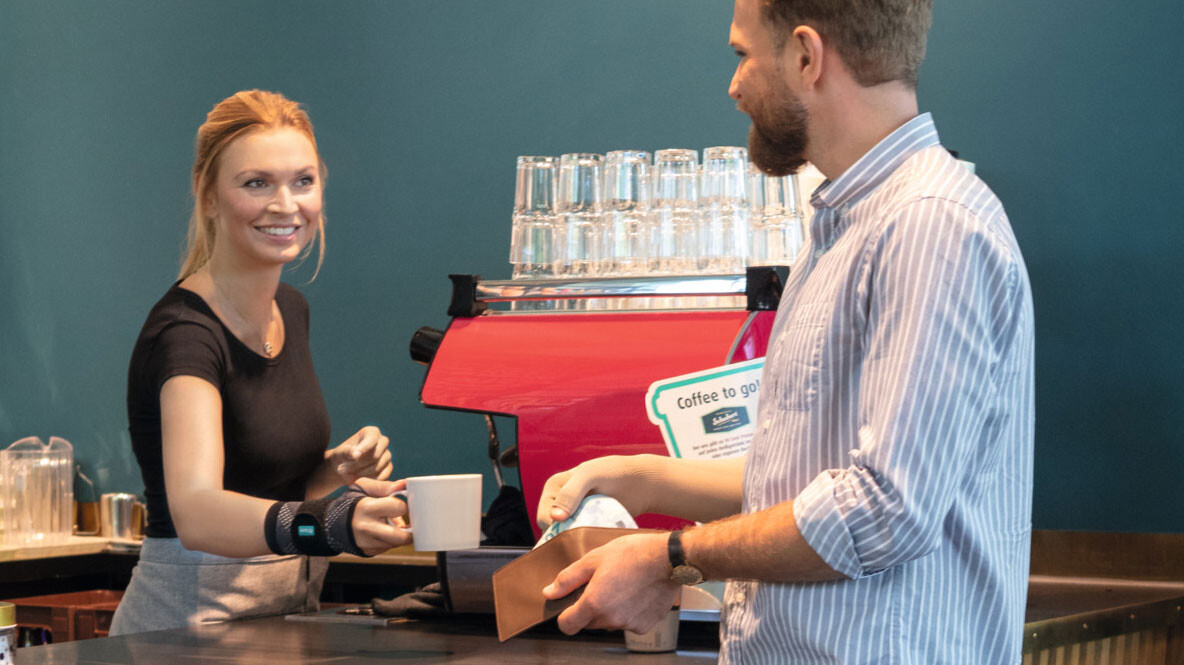 A waitress wearing a JuzoFlex Manu Xtra wrist support hands a cup to a customer over the counter A waitress wearing a JuzoFlex Manu Xtra wrist support hands a cup to a customer over the counter