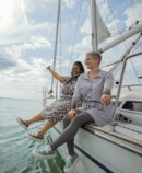 Two people are sitting relaxed at the edge of a sailing boat, looking out over the water; one is wearing compression garments on their arms and legs. Two people are sitting relaxed at the edge of a sailing boat, looking out over the water; one is wearing compression garments on their arms and legs.