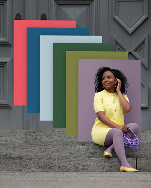 Una mujer con medias de compresión en color Cozy Violet está sentada en unas escaleras. Al fondo, los paneles de color de la actual colección Juzo Trend Colours.