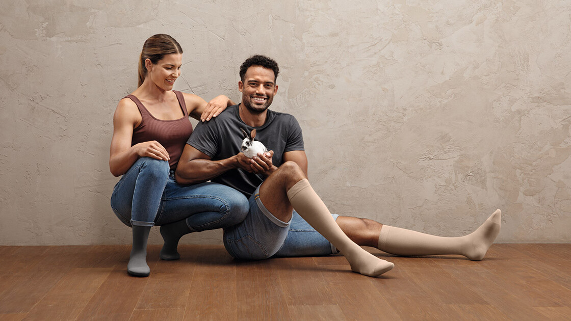 Um casal de desportistas está agachado no chão. Ambos usam meias de compressão Juzo Intenso. O homem tem um coelhinho na mão. Um casal de desportistas está agachado no chão. Ambos usam meias de compressão Juzo Intenso. O homem tem um coelhinho na mão.